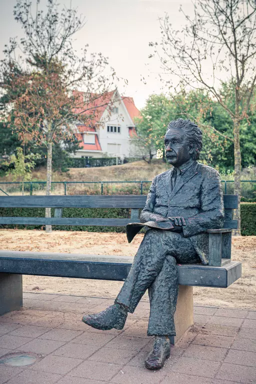 Eine Bronzeskulptur von Albert Einstein sitzt auf einer Bank, umgeben von Bäumen und einem Gebäude im Hintergrund.