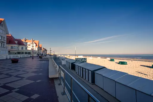 Eine Strandpromenade mit Häusern auf der linken Seite, einem blauen Himmel und einem weitläufigen Strand rechts.