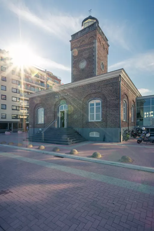 A historic brick building with a small tower, surrounded by modern structures and a cobblestone street.