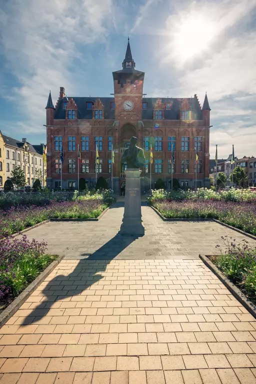 A historic red brick building with a clock, surrounded by flower beds and a statue in the foreground.