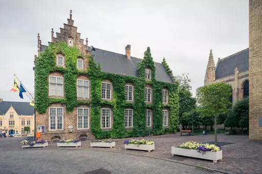 A historic building with an ivy-covered façade, surrounded by flower beds and trees in a tranquil setting.