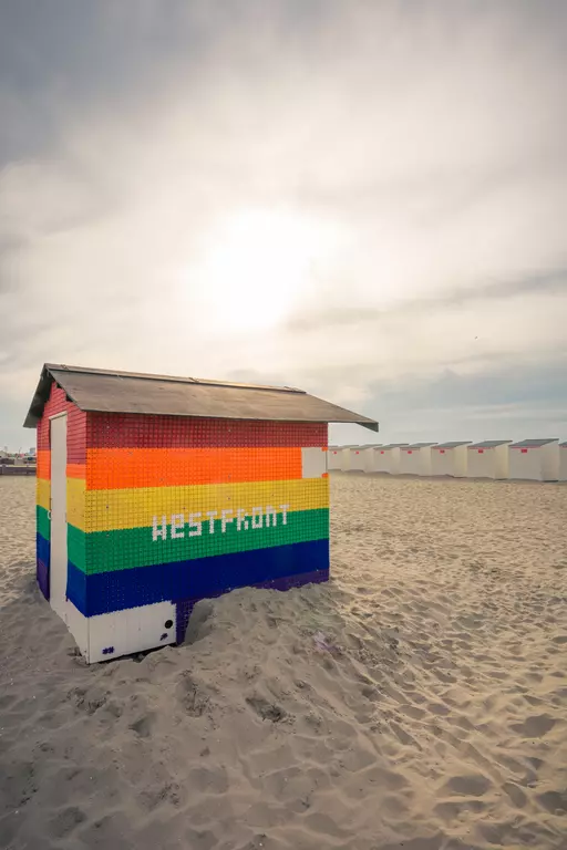 A colorful beach hut in rainbow colors stands on the sand, with additional huts visible in the background.