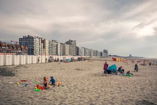 People play in the sand at the beach, surrounded by buildings and beach huts, with a cloudy sky overhead.