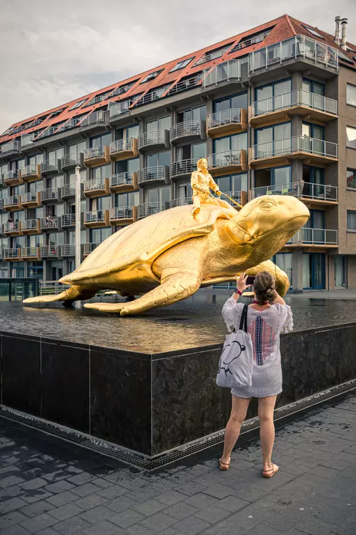 A woman in a light tunic stands in front of a large golden turtle with a figure on its back, positioned in water.