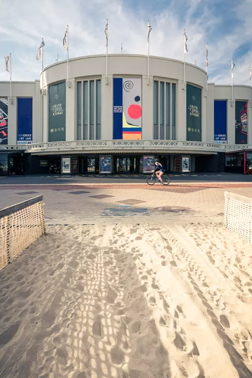 A view of a modern building with flags, colorful posters, and a sandy area in the foreground. A cyclist is passing by.