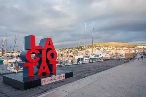 Colorful "LA CIOTAT" sign at the marina in La Ciotat, with boats and hills in the background against a cloudy sky.