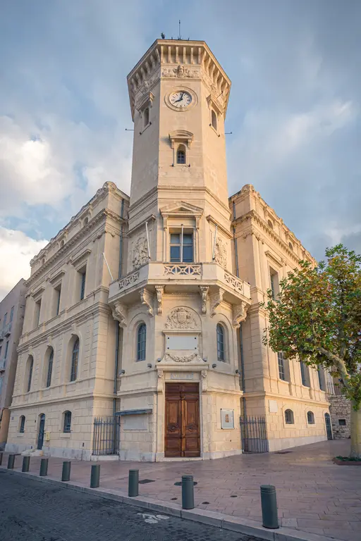 Museum building in La Ciotat, featuring a clock tower and intricate architectural details. Nearby trees and a cobblestone street add charm.