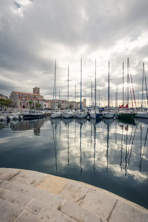 A marina in La Ciotat with sailboats reflecting in calm water under a cloudy sky, featuring a historic building in the background.