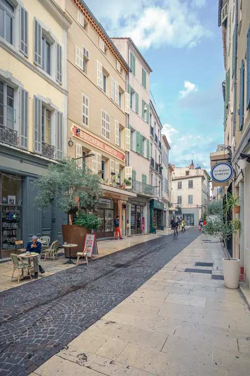 A charming street in La Ciotat lined with shops, cafes, and historic buildings under a blue sky. Cobblestone pavement adds to the ambiance.