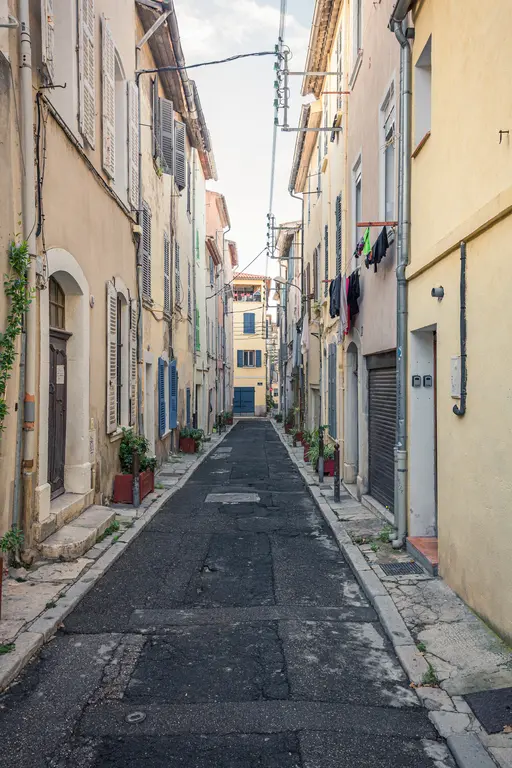A narrow street in La Ciotat lined with rustic buildings, blue window shutters, and potted plants on either side.