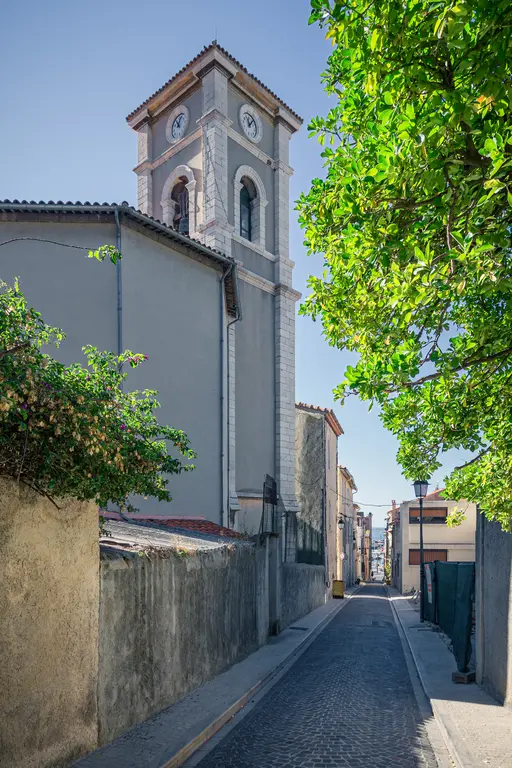 Straße in Cassis mit der Église Saint-Michel im Hintergrund. Die schmalen Häuser flankieren die gepflasterte Straße, umgeben von Bäumen.