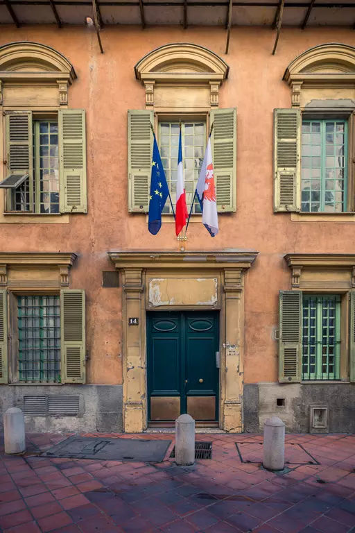 An orange building facade with green shutters, three flags, and a dark entrance door.