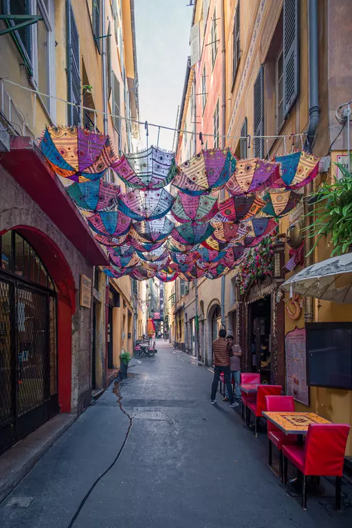 Colorful umbrellas hang over a narrow alley lined with cafes and shops, displaying cheerful colors and plants.