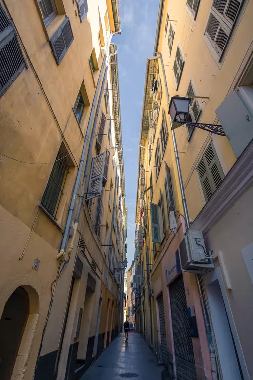 Narrow alleyway between tall yellow buildings with windows and hanging laundry. A person stands in the center of the street.