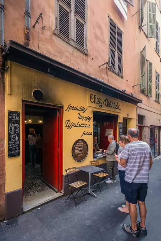 A small bakery with a yellow facade and red entrance, outdoor tables, with guests waiting in line.