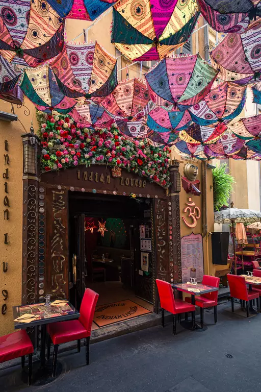 Colorful umbrellas hang above the entrance of an Indian restaurant, surrounded by red tables and flowers.