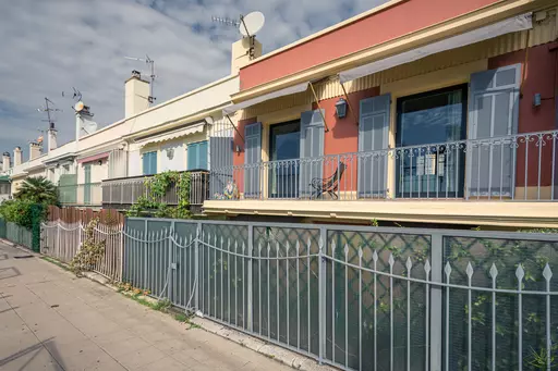 A row of houses with balconies, window shutters, and a decorative metal fence, surrounded by a paved walkway and greenery.