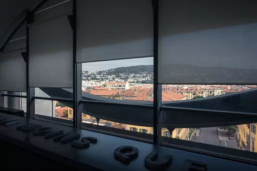 View through a window with shades overlooking red rooftops and hills in the background.