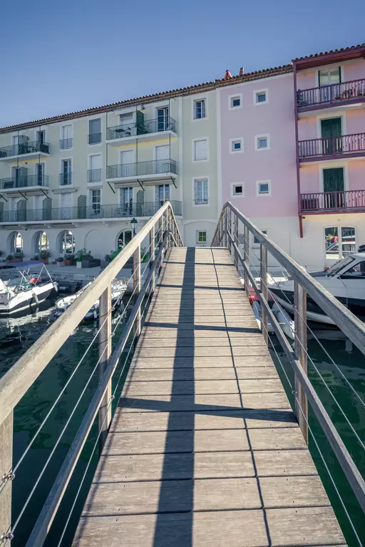 Ein Holzsteg führt über Wasser zu bunten Wohnhäusern, die an einem Hafen stehen, mit Booten, die docken.