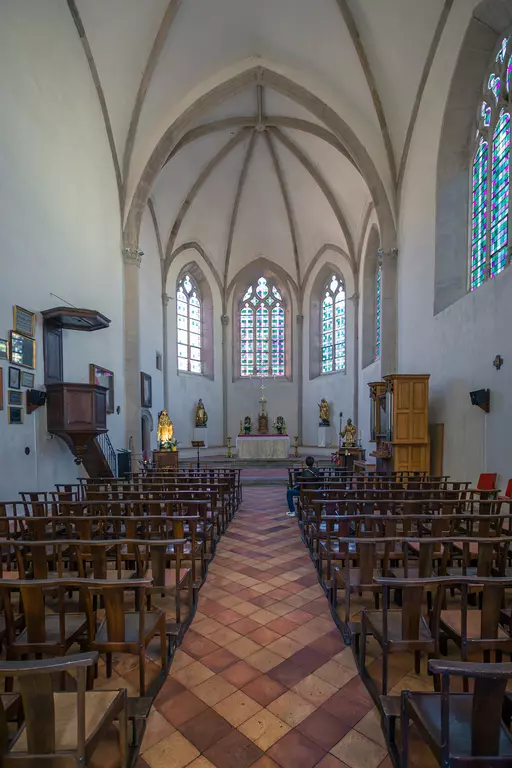 Helle Kirche mit hohen, gewölbten Decken, Holzstühlen in Reihen, bunten Fenstern und einem Altar mit Statuen im Hintergrund.