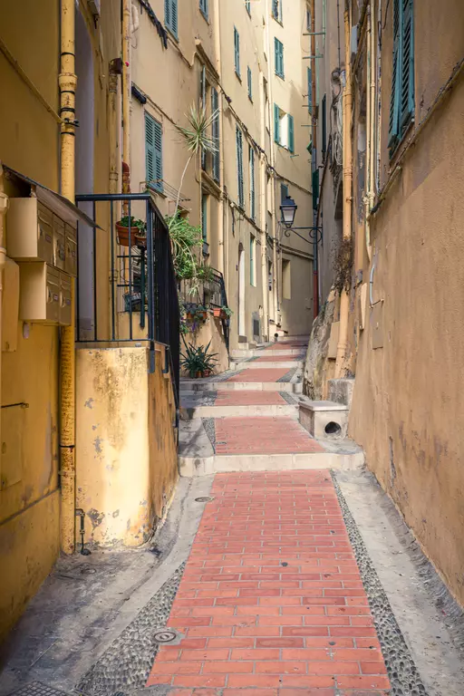 A narrow, cobbled path between tall yellow buildings with shutters and plants. A lantern hangs on the wall.