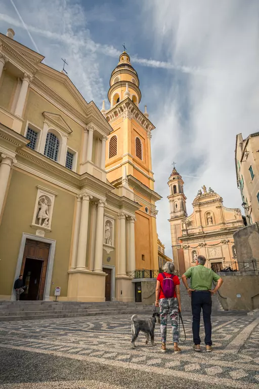 A woman and a man stand with a dog in a town featuring historic buildings and a tall church tower in the background.