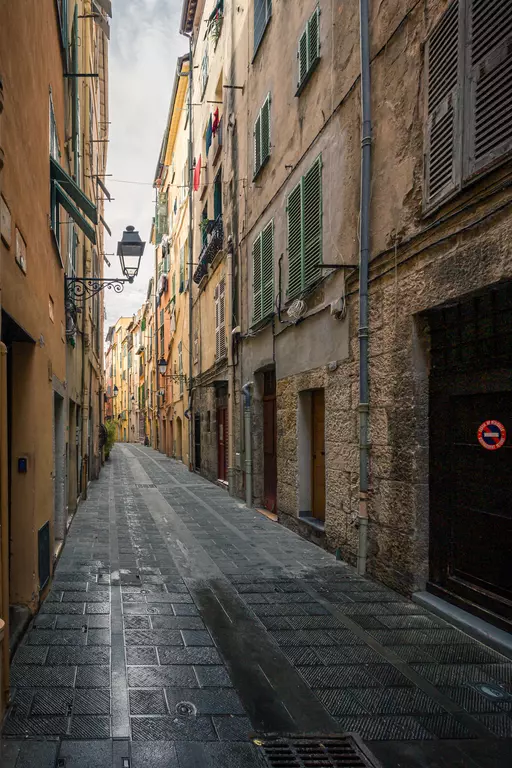 A narrow, cobbled street with old buildings, green-shuttered windows, and a streetlamp in the background.