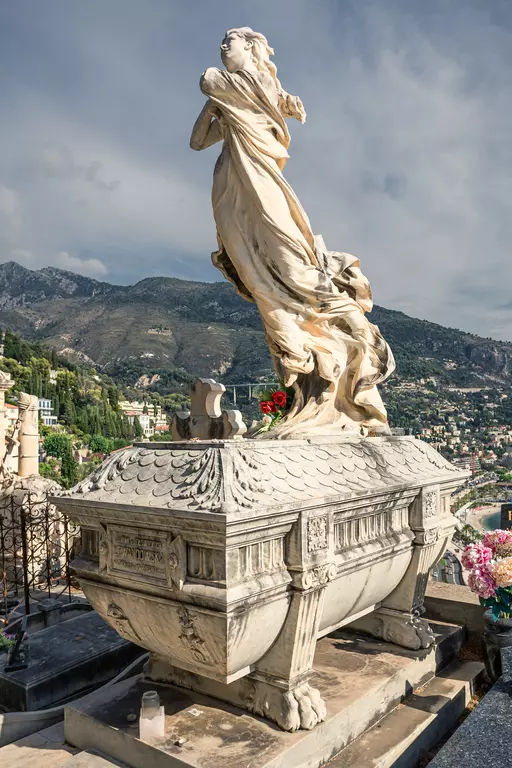 A statue of a woman in flowing garments stands on a decorated grave. Mountains and a cityscape are visible in the background.