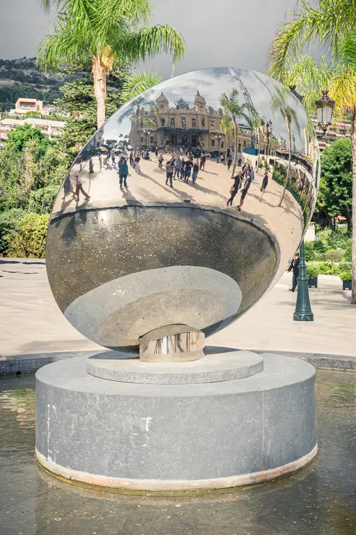 A large, shiny oval sculpture stands on a round base, surrounded by people and green palm trees.