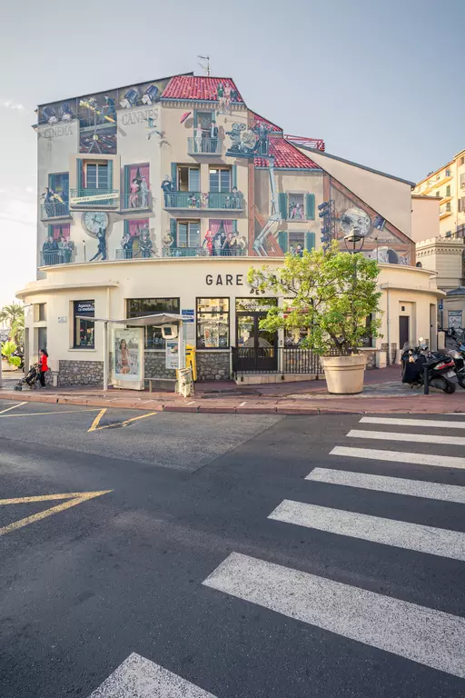 Bunt bemaltes Gebäude mit dem Schriftzug GARE. Menschen gehen über Zebrastreifen. Im Vordergrund ein Blumentopf, im Hintergrund Motorräder.