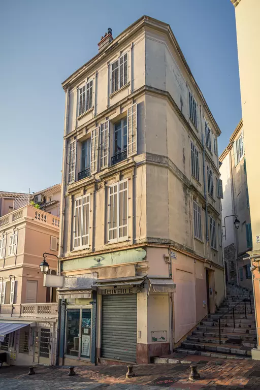 Ein dreigeschossiges, gelbes Gebäude mit weißen Fensterläden und einer geschlossenen Ladenfront. Treppen führen zur Seite des Hauses.