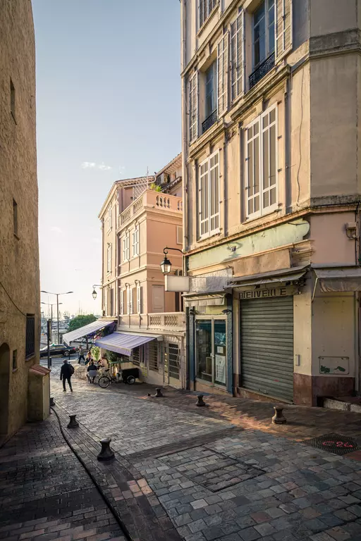 Gasse mit gepflastertem Boden, umgeben von pastellfarbenen Gebäuden und einem kleinen Marktstand. Eine Person geht vorbei.