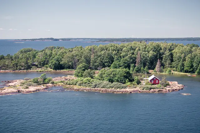 Kleine Insel im Wasser mit dichtem Grün und einem roten Holzhaus. Steinige Küstenlinie und ruhige Wasseroberfläche im Hintergrund.