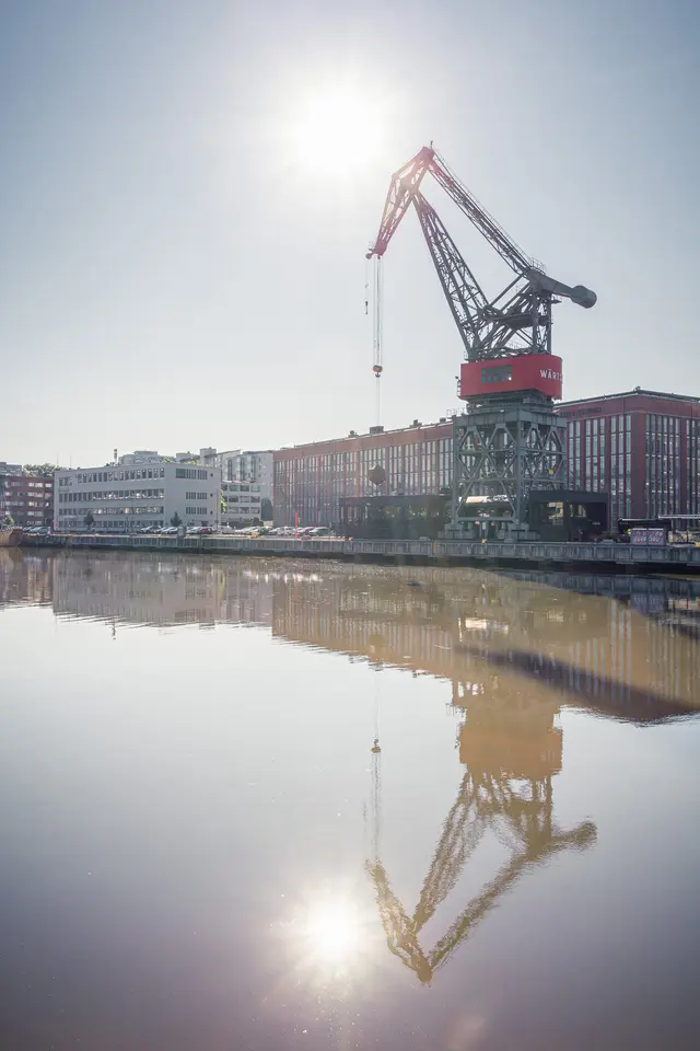 Hafenkran spiegelt sich im ruhigen Wasser, umgeben von modernen Gebäuden und bläulichem Himmel, mit der Sonne im Hintergrund.