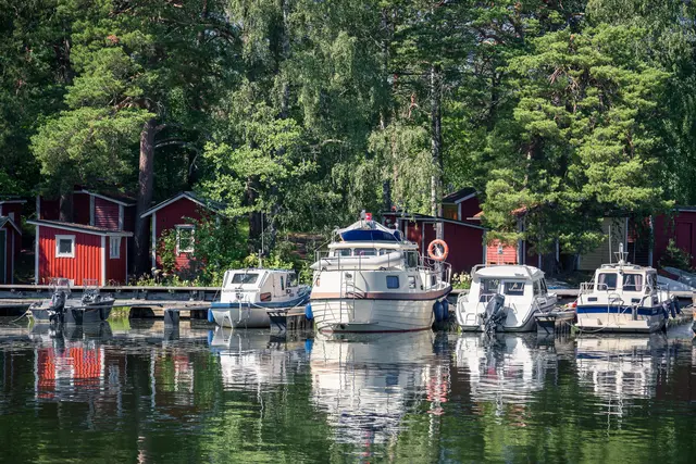 Boote am Steg in ruhigem Wasser, umgeben von grünen Bäumen und roten Holzgebäuden. Die Spiegelungen im Wasser sind deutlich zu sehen.
