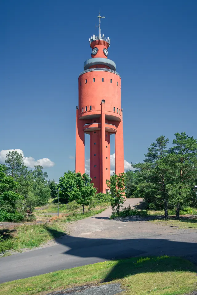 Roter Wasserturm mit markanter Form, umgeben von Bäumen und Wiese. Ein blauer Himmel bildet den Hintergrund.