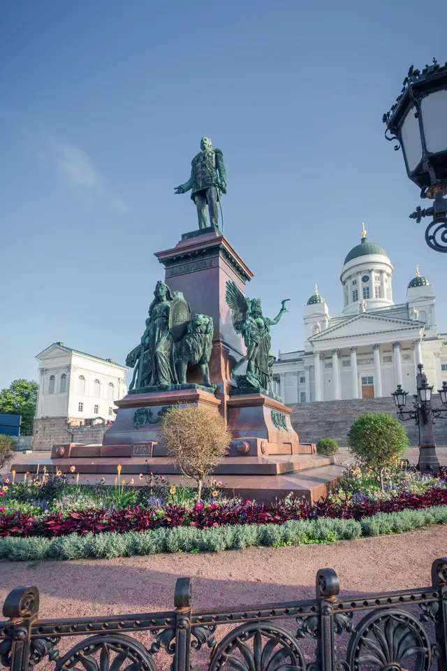 A bronze monument featuring historical figures, surrounded by colorful flowers and a decorative fence, with an impressive building in the background.