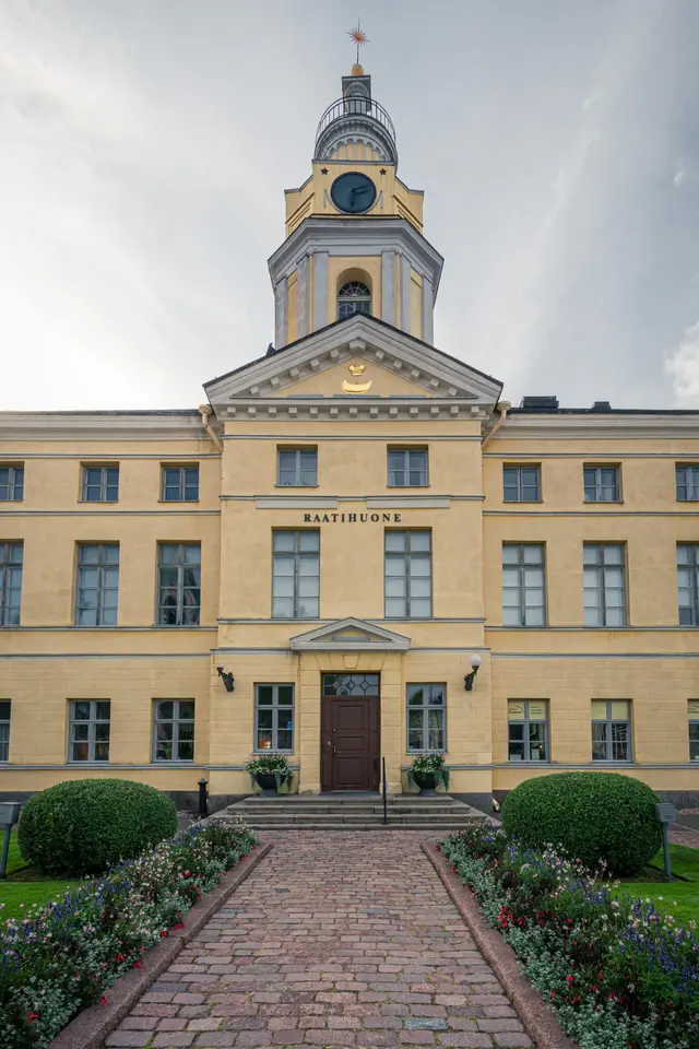 Rathaus mit gelber Fassade und Uhrturm, umgeben von einem gepflasterten Weg und grünen Sträuchern. Blumenbeete an den Seiten.