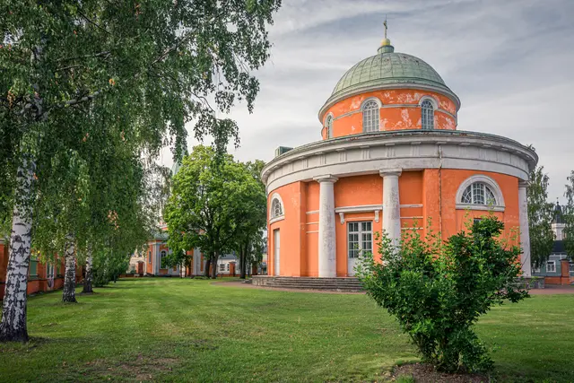 Runder, orangefarbener Gebäudekomplex mit Kuppeldach und Säulen in einer grünen, gepflegten Landschaft, umgeben von Bäumen.