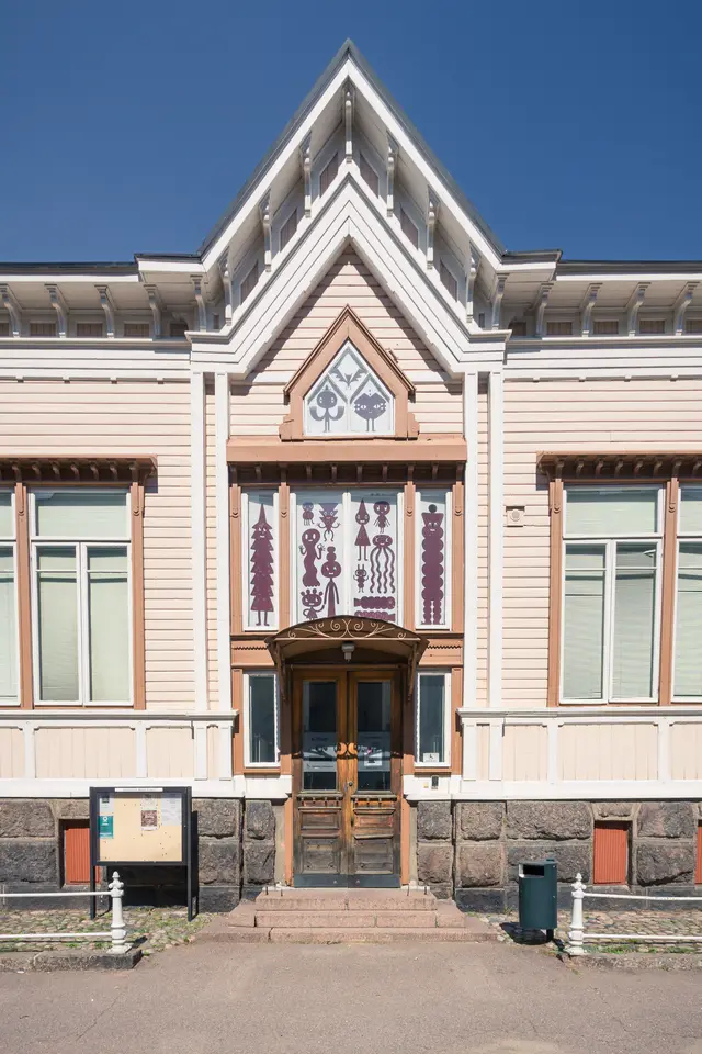 Historisches Gebäude mit Holzfassade, verzierten Fenstern und doppelflügeliger Eingangstür. Dunkler Sockel und Informationsschilder befinden sich im Vordergrund.