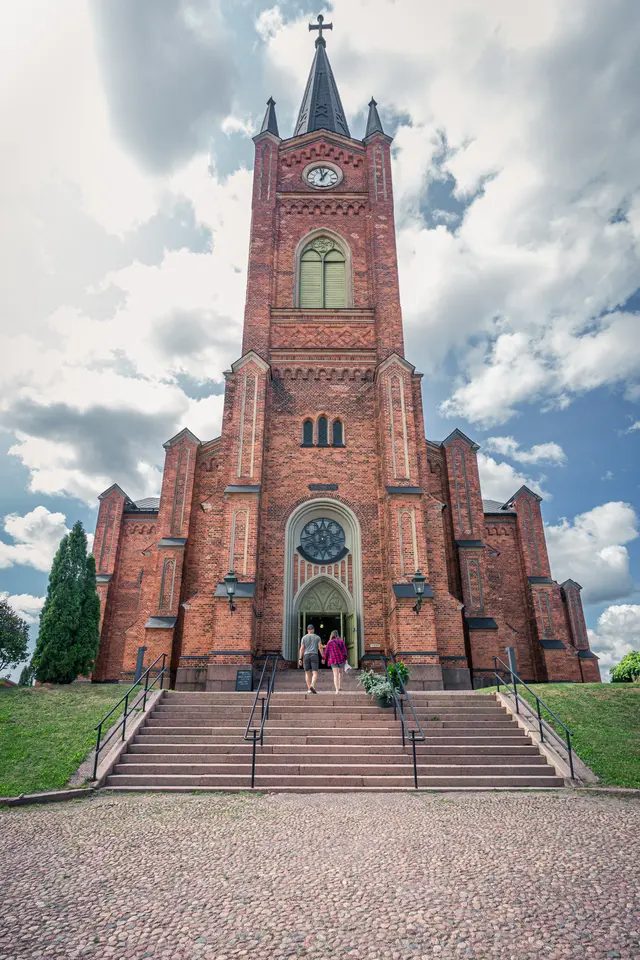Historische Kirche aus roten Ziegeln mit hohem Turm, umgeben von Stufen, auf denen zwei Personen zur Eingangstür gehen.