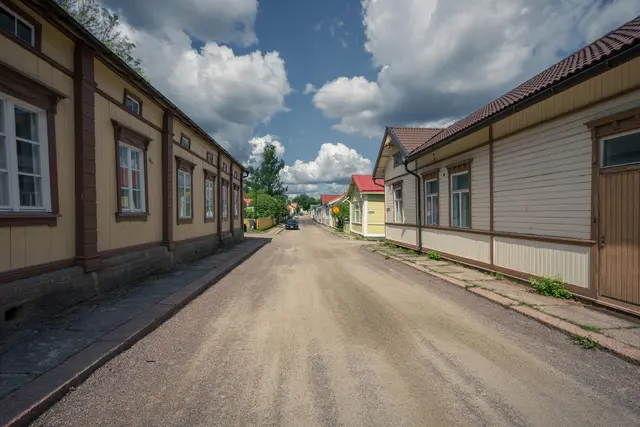 Blick auf eine schmale, sandige Straße mit bunten Holzhäusern auf beiden Seiten. Über dem Bild ziehen weiße Wolken am blauen Himmel.