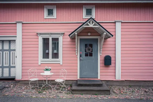 Haus mit rosa Holzfassade, grau-blauer Eingangstür und geschmücktem Eingangsbereich. Neben der Tür stehen ein kleiner Tisch und zwei Stühle.