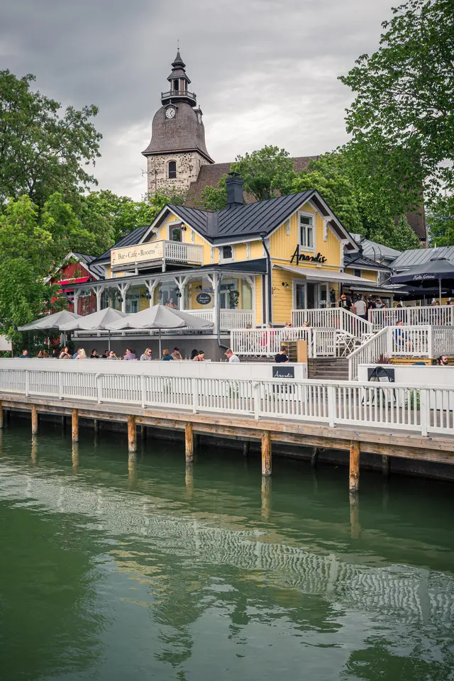 Blick auf ein gelbes Café mit Terrasse am Wasser, umgeben von Bäumen und einem alten Turm im Hintergrund. Gastgäste sitzen auf der Terrasse.