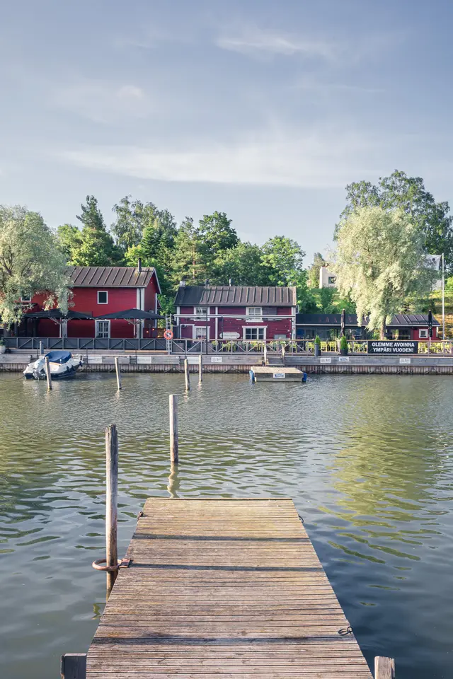 Holzsteg am Wasser mit rot gestrichenen Häusern im Hintergrund, umgeben von Bäumen und sanften Wellen.