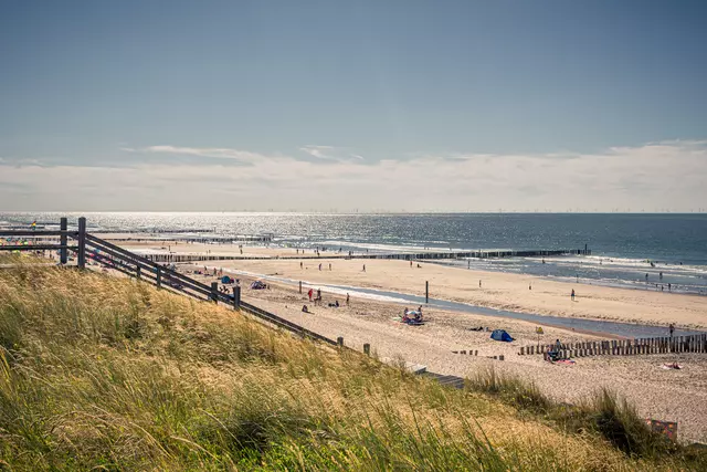 Der Sandstrand erstreckt sich bis zum Wasser, mit einigen Menschen, die sich entspannen. Wellen brechen sanft am Ufer, und im Hintergrund sind Stege sichtbar.