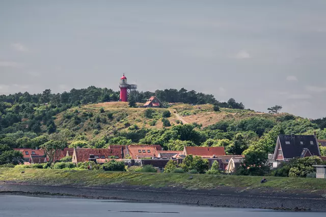 Leuchtturm auf Vlieland: Ein roter Leuchtturm thront auf einem grünen Hügel über farbenfrohe Häuser am Ufer.