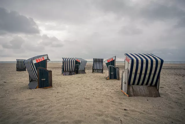 Sechs Strandkörbe mit blauen und weißen Streifen stehen im Sand unter einem bewölkten Himmel.