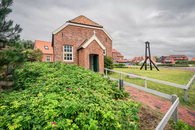 Ein kleines Backsteingebäude mit einem Satteldach und einem Kreuz, umgeben von grünen Pflanzen und einer kleinen Wiese.