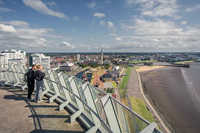 Zwei Personen stehen an einem Geländer mit Blick auf eine Küstenstadt, umgeben von Wasser, Stränden und Wolken.